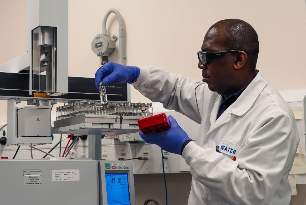 Person in a lab coat and blue gloves conducting water quality testing, the person is holding a small vial in their right hand and red device in left hand. 