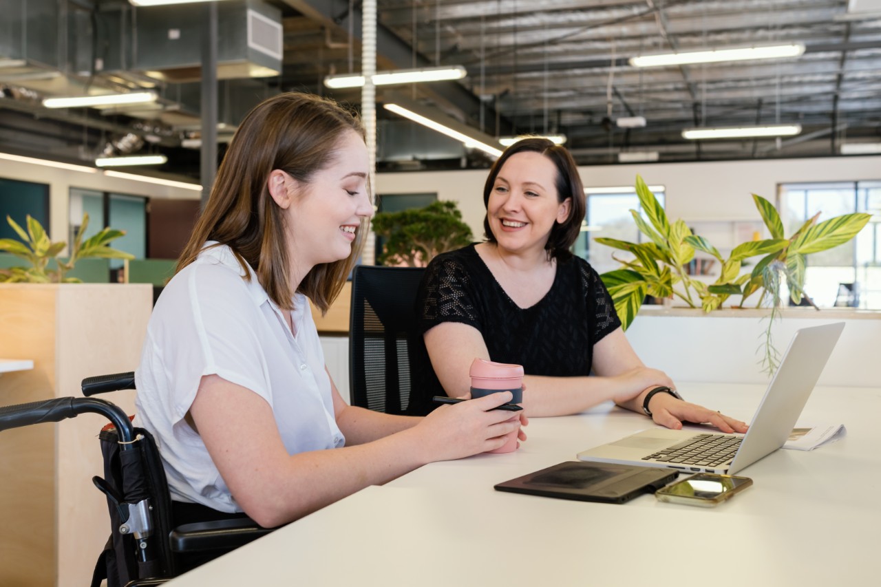 Two female Sydney Water employees, one in a wheelchair, collaborating at a desk with laptops. 
