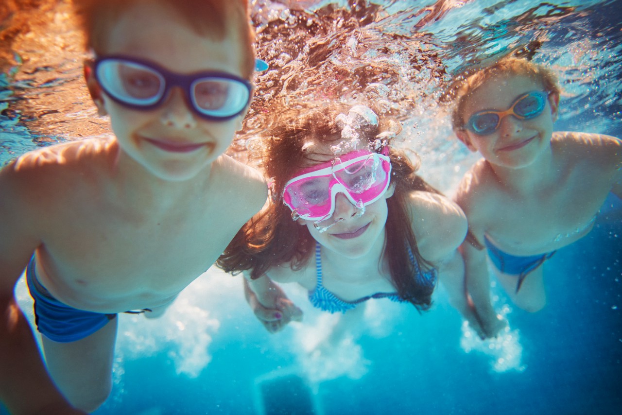 A shot of three children underwater, wearing goggles and holding hands while smiling at the camera. 