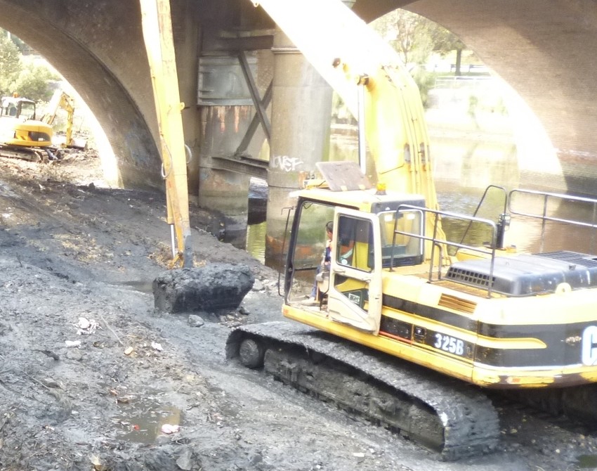 A large excavator scooping up silt next to river, showing the Cooks River silt removal process