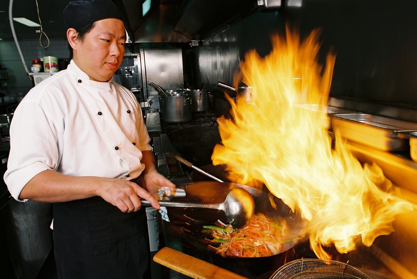 A chef stir fries noodles and vegetables on a stove.