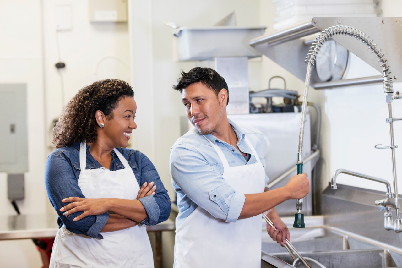 A man and woman in a commercial kitchen, the man holding a spray tap in the sink. 