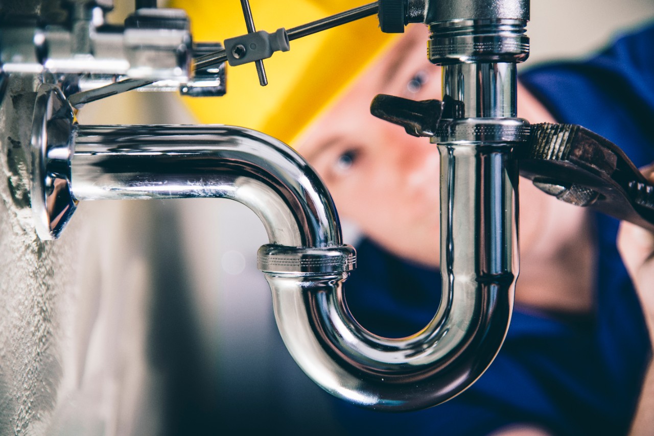Closeup photo of drain pipes, with a plumber behind it. He is using a wrench to tighten a valve. 