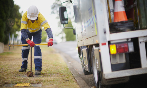 A Sydney Water crew worker access a hydrant using a special tool, with a Sydney Water truck parked next to him. 